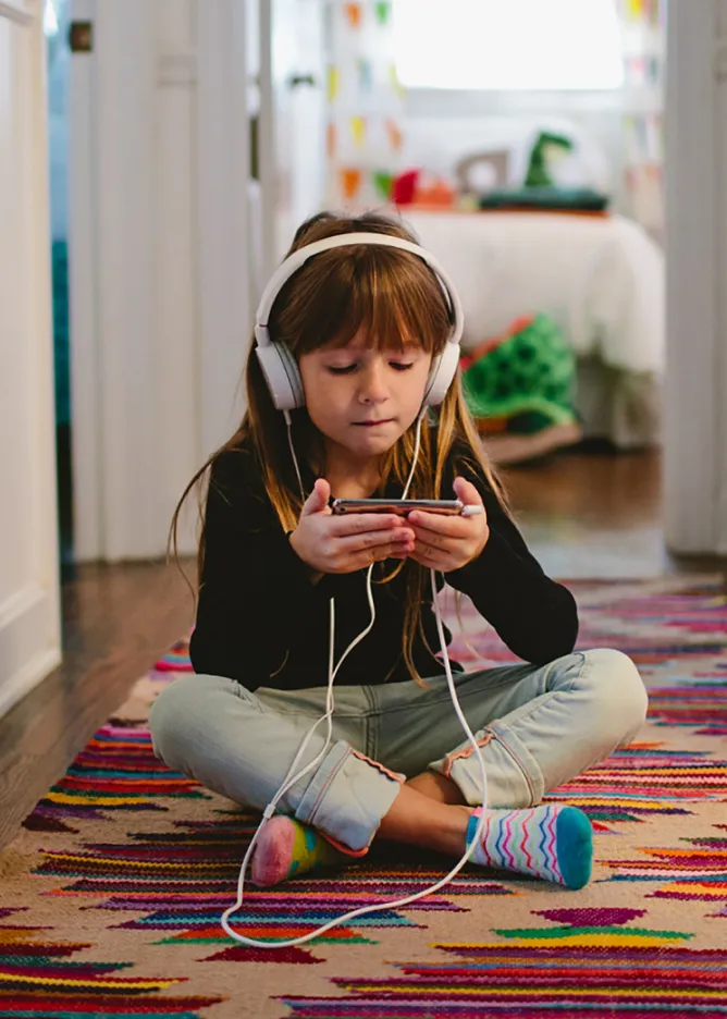 Child sitting cross-legged on the floor — using her phone and headphones